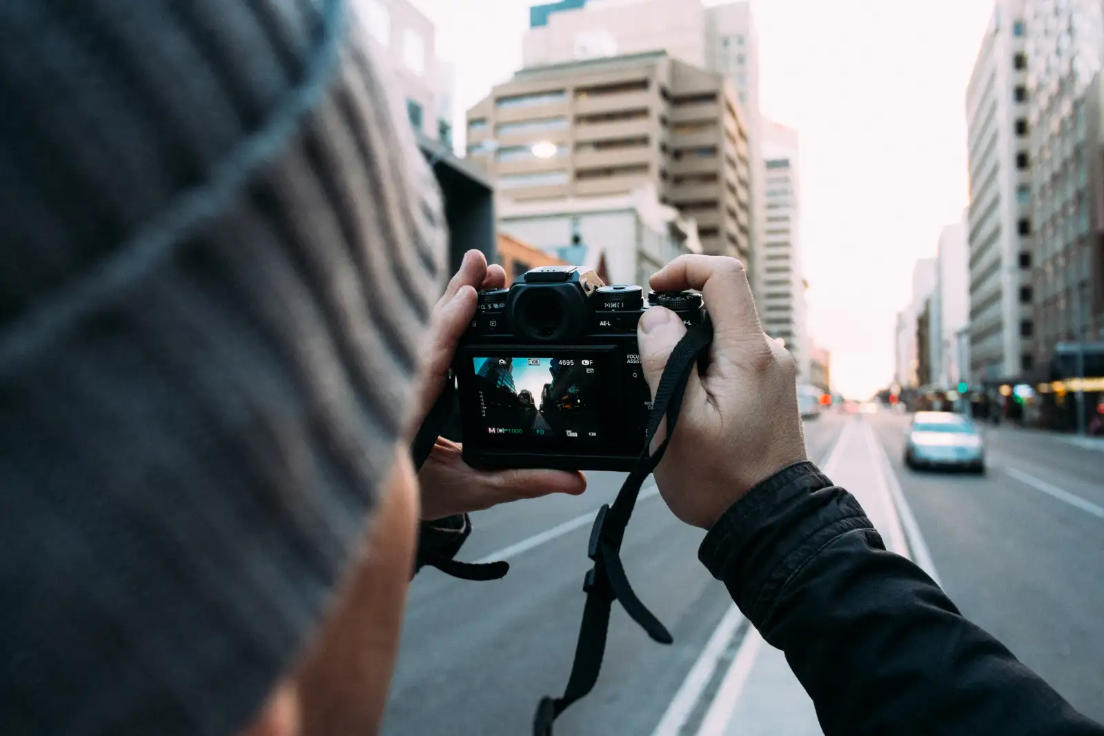 Two hands holding a mirrorless camera, its screen showing a city street