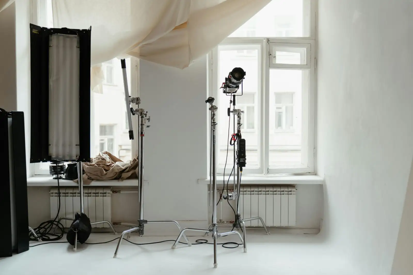 A white-walled daylight studio with a large window, a light stand and a hanging diffusion scrim